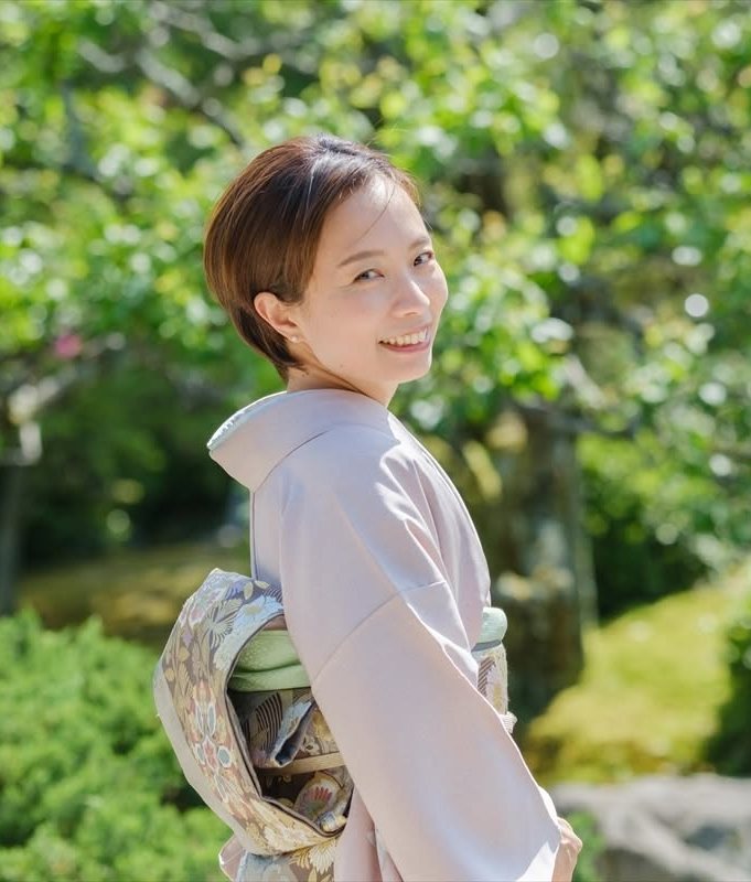 ナビゲーター 古原治奈。淡い藤色の着物で庭園に立ち、ふり返って微笑む姿